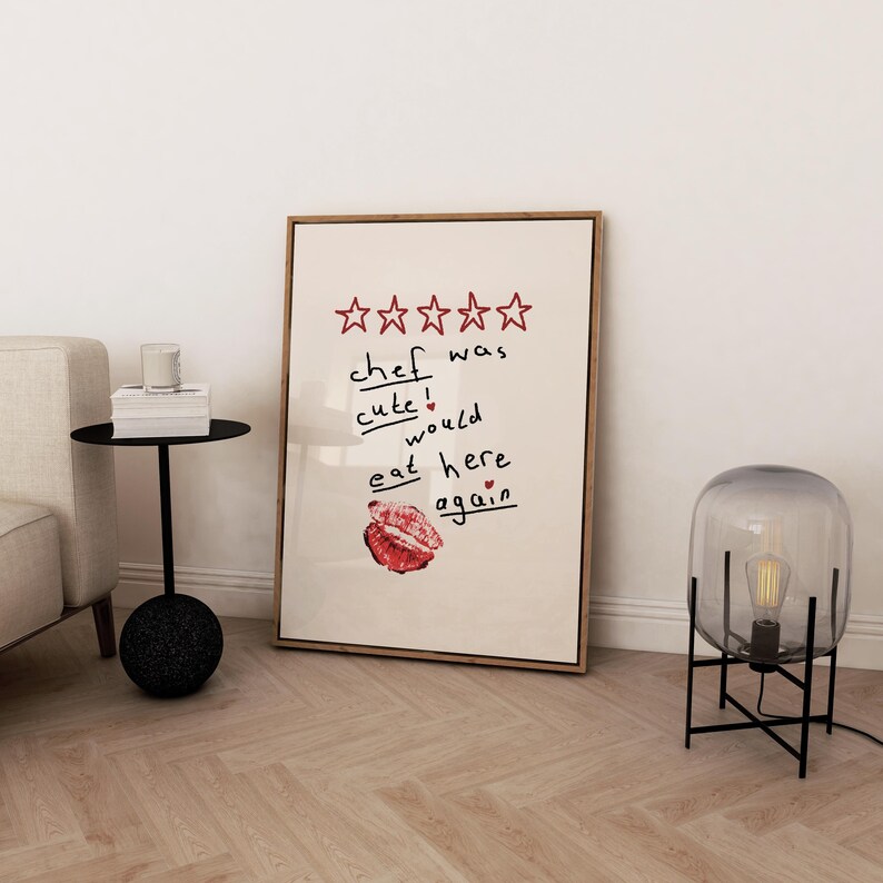Op de afbeelding: Een ingelijst print met een vijfsterrenbeoordeling en de tekst "kok was schattig! zou hier weer eten". De print hangt aan een witte muur boven een zwarte bijzettafel met een glazen kaarsenhouder en boeken.