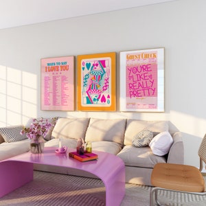 May include: Three framed prints on a white wall above a beige sofa. The prints are all in a retro style with pink, blue, and yellow colors. The left print says "Ways to Say I Love You" and has a list of phrases. The middle print is a queen of hearts playing card with a woman in a crown and sunglasses. The right print says "Guest Check" and has the text "You're Like Really Pretty" in pink and blue.