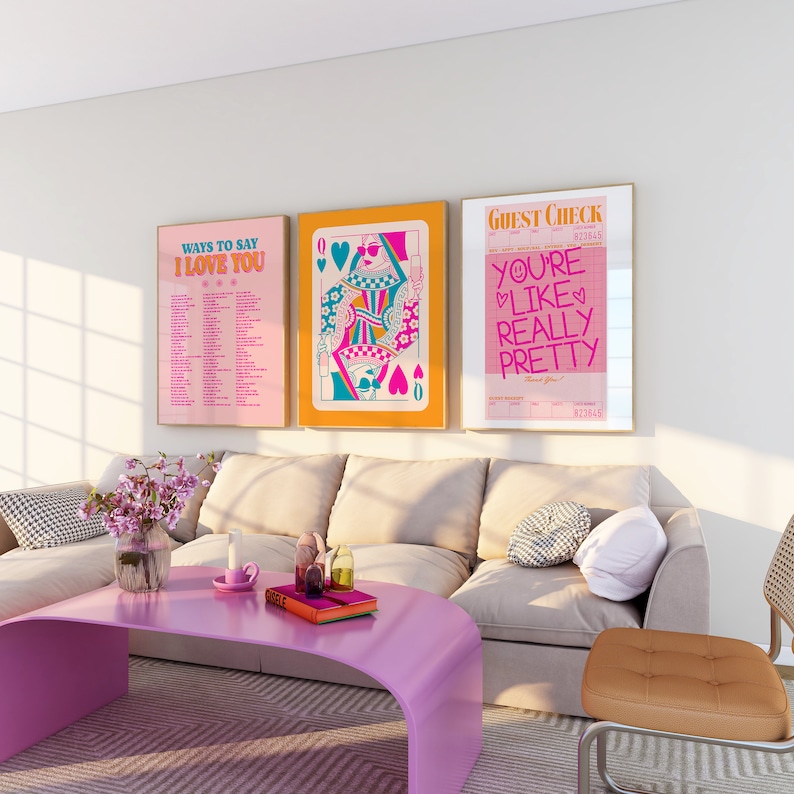 May include: Three framed prints on a white wall above a beige sofa. The prints are all in a retro style with pink, blue, and yellow colors. The left print says "Ways to Say I Love You" and has a list of phrases. The middle print is a queen of hearts playing card with a woman in a crown and sunglasses. The right print says "Guest Check" and has the text "You're Like Really Pretty" in pink and blue.
