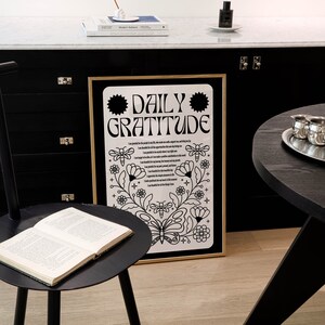 May include: A black and white framed print with the text "DAILY GRATITUDE" and a floral design. The print is hanging on a black wall with a black table and a book on a black side table.
