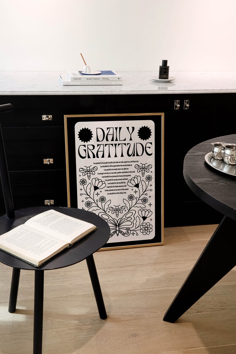 May include: A black and white framed print with the text "DAILY GRATITUDE" and a floral design. The print is hanging on a black wall with a black table and a book on a black side table.