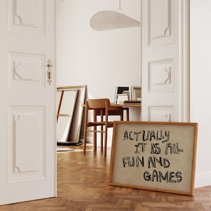 Pode incluir: Uma placa emoldurada com o texto "ACTUALLY IT IS ALL FUN AND GAMES" em letras pretas. A placa est&aacute; encostada em um piso de madeira em um quarto com paredes brancas e uma porta parcialmente aberta.