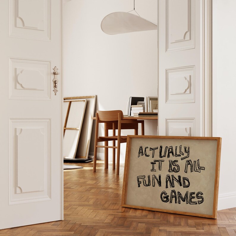 Pode incluir: Uma placa emoldurada com o texto "ACTUALLY IT IS ALL FUN AND GAMES" em letras pretas. A placa est&aacute; encostada em um piso de madeira em um quarto com paredes brancas e uma porta parcialmente aberta.