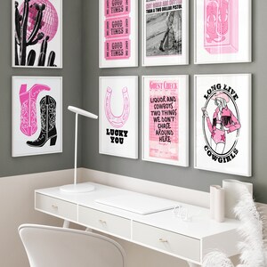 May include: A collection of eight pink and black framed prints with various designs, including a disco ball, a horseshoe, a cowboy boot, a guest check, and a cowgirl. The prints are hanging on a gray wall above a white desk with a white chair.