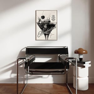 May include: Black and white illustration of a martini glass with a cherry and a splash of liquid. The text "Manhattan" is written in a stylized font below the glass. The image is framed and hanging on a white wall.