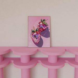 May include: A framed print of two pink disco ball strawberries on a pink background. The print is hanging on a wall above a pink shelf.