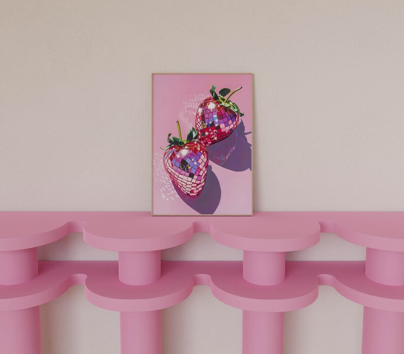 May include: A framed print of two pink disco ball strawberries on a pink background. The print is hanging on a wall above a pink shelf.