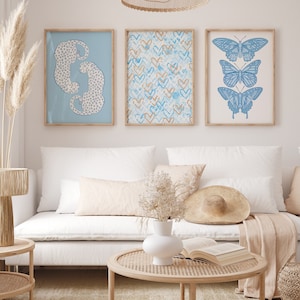 May include: Three framed prints on a white wall above a white couch. The prints feature a blue and white leopard print, a blue and gold heart pattern, and a blue butterfly illustration.