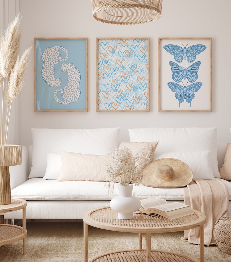 May include: Three framed prints on a white wall above a white couch. The prints feature a blue and white leopard print, a blue and gold heart pattern, and a blue butterfly illustration.