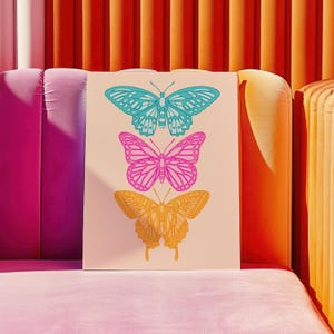 Pode incluir: Uma impress&atilde;o com tr&ecirc;s borboletas estilizadas em uma linha vertical. A borboleta superior &eacute; azul, a borboleta do meio &eacute; rosa e a borboleta inferior &eacute; amarela.
