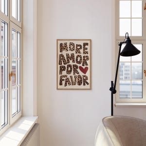 May include: A framed print with the text "MORE AMOR POR FAVOR" in a leopard print font with a red heart in the middle of the text. The print is hanging on a white wall.