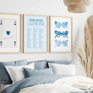 May include: Three framed prints on a white wall above a bed with blue bedding. The left print is a playing card, the ace of hearts. The middle print has a list of ways to say "I love you". The right print has three pairs of blue heart-shaped sunglasses.