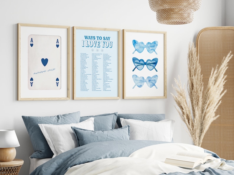 May include: Three framed prints on a white wall above a bed with blue bedding. The left print is a playing card, the ace of hearts. The middle print has a list of ways to say "I love you". The right print has three pairs of blue heart-shaped sunglasses.