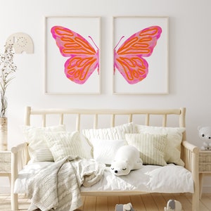 May include: Two framed prints of a pink and orange butterfly with a white background. The prints are hanging on a white wall above a white couch with pillows and a blanket.