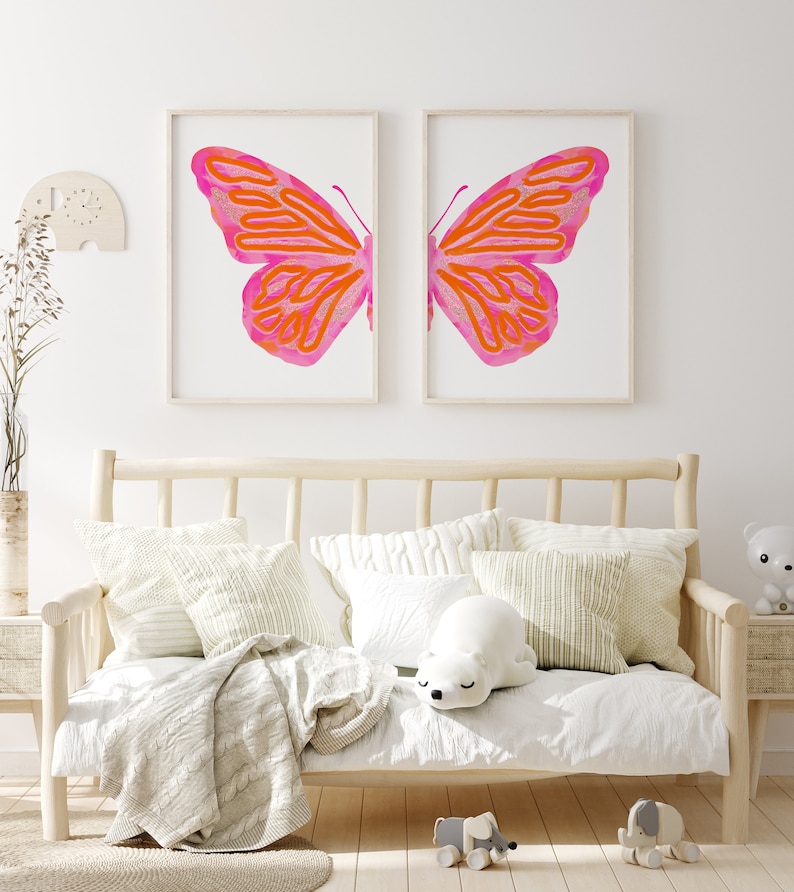 May include: Two framed prints of a pink and orange butterfly with a white background. The prints are hanging on a white wall above a white couch with pillows and a blanket.