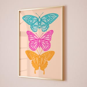 Pode incluir: Uma impress&atilde;o emoldurada com tr&ecirc;s borboletas dispostas verticalmente. A borboleta superior &eacute; azul, a borboleta do meio &eacute; rosa e a borboleta inferior &eacute; amarela. As borboletas s&atilde;o delineadas em preto e t&ecirc;m um fundo branco.
