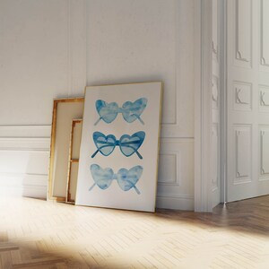 May include: A framed print of three watercolor illustrations of blue heart-shaped sunglasses. The sunglasses are arranged vertically in a row.