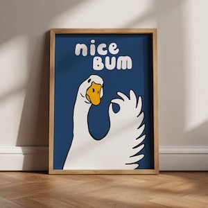 May include: A framed art print featuring a cartoon goose with a yellow beak and a white body, giving an "okay" hand gesture. The words "nice bum" are written in white, bubble letters against a dark blue background.