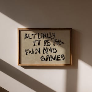 Pode incluir: Uma impress&atilde;o emoldurada com o texto "ACTUALLY IT IS ALL FUN AND GAMES" em letras pretas, estilo manuscrito. A impress&atilde;o tem um fundo bege e est&aacute; emoldurada em uma moldura de madeira marrom clara. A imagem &eacute; exibida em uma parede branca.