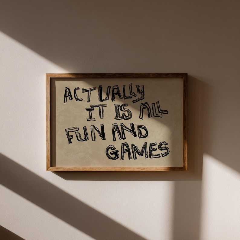 Pode incluir: Uma impress&atilde;o emoldurada com o texto "ACTUALLY IT IS ALL FUN AND GAMES" em letras pretas, estilo manuscrito. A impress&atilde;o tem um fundo bege e est&aacute; emoldurada em uma moldura de madeira marrom clara. A imagem &eacute; exibida em uma parede branca.