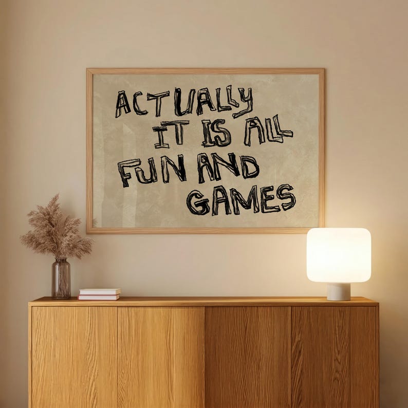 Pode incluir: Uma impress&atilde;o emoldurada com o texto "ACTUALLY IT IS ALL FUN AND GAMES" em letras pretas. A impress&atilde;o &eacute; exibida acima de um arm&aacute;rio de madeira, com uma l&acirc;mpada e um vaso decorativo.