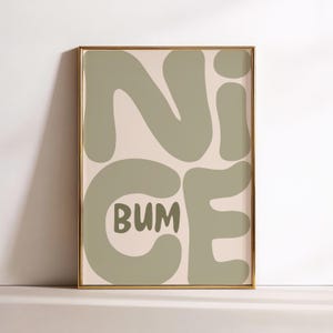 May include: A framed art print with the words "NICE BUM" in a playful, rounded font. The letters are a muted green against a light pink background. The frame is gold.