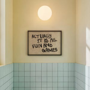 Pode incluir: Uma obra de arte emoldurada com o texto "ACTUALLY IT IS ALL FUN AND GAMES" est&aacute; pendurada na parede. A parede &eacute; pintada em amarelo claro, e uma lumin&aacute;ria redonda e branca &eacute; montada acima da obra de arte. Abaixo da obra, a parede &eacute; revestida em azul claro.
