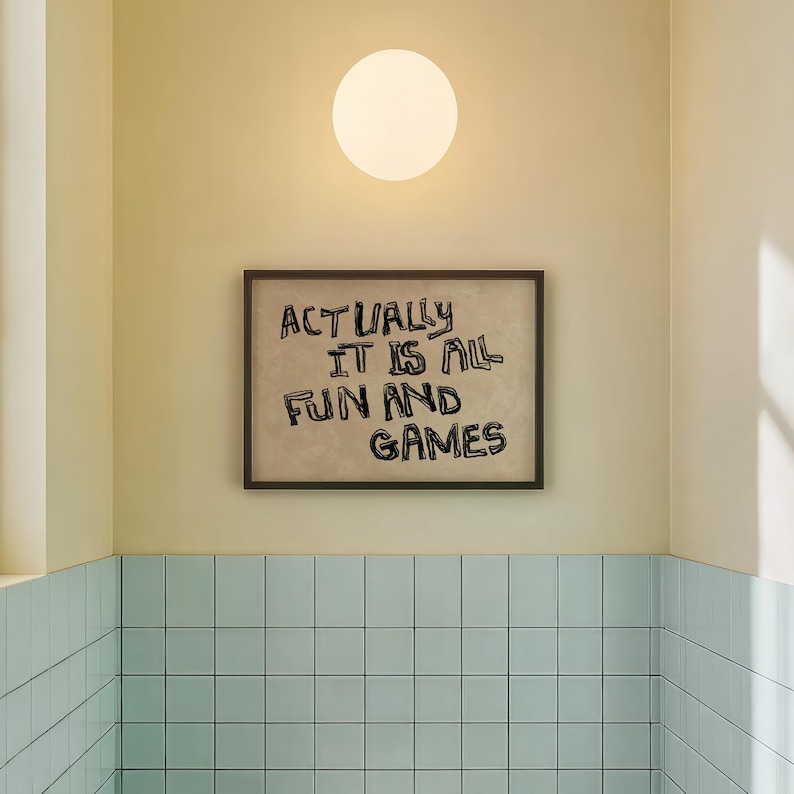Pode incluir: Uma obra de arte emoldurada com o texto "ACTUALLY IT IS ALL FUN AND GAMES" est&aacute; pendurada na parede. A parede &eacute; pintada em amarelo claro, e uma lumin&aacute;ria redonda e branca &eacute; montada acima da obra de arte. Abaixo da obra, a parede &eacute; revestida em azul claro.