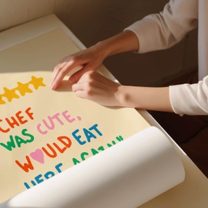 May include: A yellow poster with the text "Chef was cute, would eat here again" written in colorful letters. Five yellow stars are at the top of the poster. A person's hand is rolling up the poster.