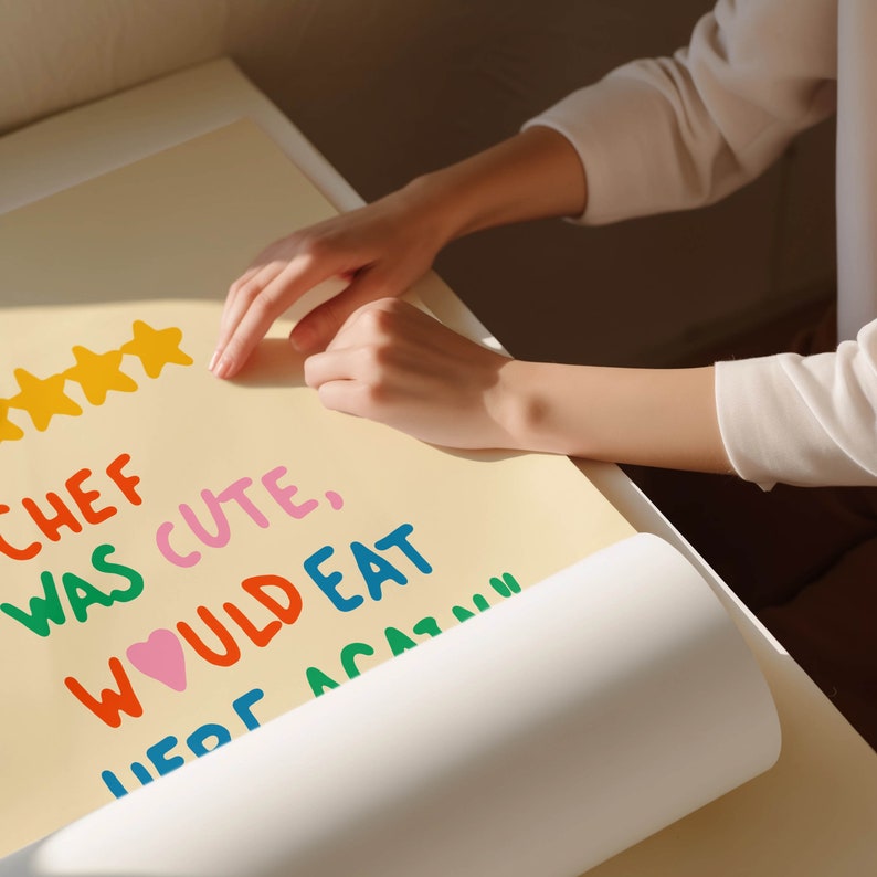 May include: A yellow poster with the text "Chef was cute, would eat here again" written in colorful letters. Five yellow stars are at the top of the poster. A person's hand is rolling up the poster.
