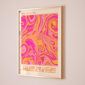 Peut inclure: Une impression encadr&eacute;e avec un motif abstrait en spirale rose, orange et jaune. Le texte "Trust the Universe" est en haut et "You are the luckiest person in the world" est en bas.