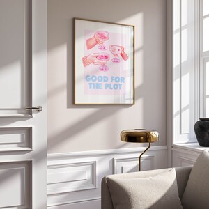 May include: A gold framed print with a pink background and an illustration of four hands holding up glasses of pink champagne. The text "GOOD FOR THE PLOT" is printed in blue letters below the illustration.