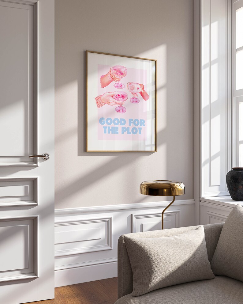 May include: A gold framed print with a pink background and an illustration of four hands holding up glasses of pink champagne. The text "GOOD FOR THE PLOT" is printed in blue letters below the illustration.