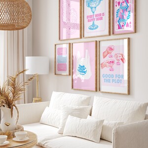 Puede incluir: Un interior de sala de estar con un sof&aacute; blanco y almohadas, una mesa de caf&eacute; de madera y una l&aacute;mpara. La pared est&aacute; decorada con obras de arte enmarcadas que presentan ilustraciones rosas y azules con texto como "Every Hour is Happy Hour", "Good Times", "Thank You for Coming!" y "Good for the Plot".