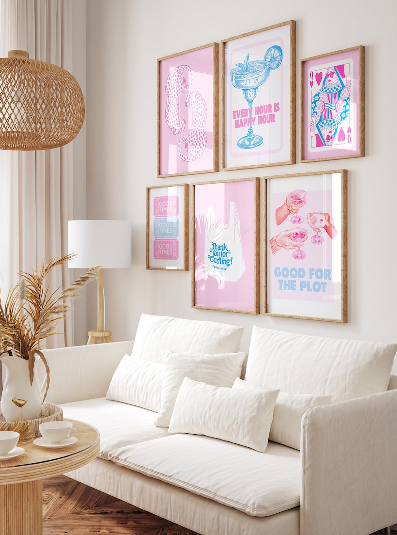 Puede incluir: Un interior de sala de estar con un sof&aacute; blanco y almohadas, una mesa de caf&eacute; de madera y una l&aacute;mpara. La pared est&aacute; decorada con obras de arte enmarcadas que presentan ilustraciones rosas y azules con texto como "Every Hour is Happy Hour", "Good Times", "Thank You for Coming!" y "Good for the Plot".