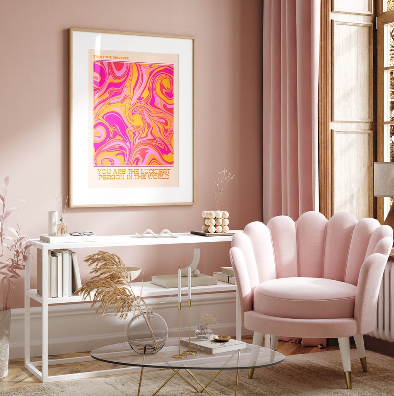 Peut inclure: Une impression abstraite en spirale rose et jaune avec le texte "You are the luckiest person in the world" en police d'or. L'impression est encadr&eacute;e dans un cadre dor&eacute; et accroch&eacute;e &agrave; un mur rose. Un fauteuil en velours rose &agrave; dossier festonn&eacute; est au premier plan.