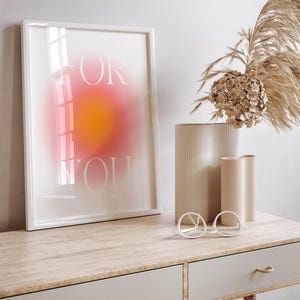 Peut inclure: Une impression encadr&eacute;e blanche avec les mots "FOR YOU" en police blanche sans empattement sur un fond d&eacute;grad&eacute; rose et orange. L'impression est pos&eacute;e sur un bureau en bois clair avec deux tiroirs et une paire de lunettes rondes blanches.
