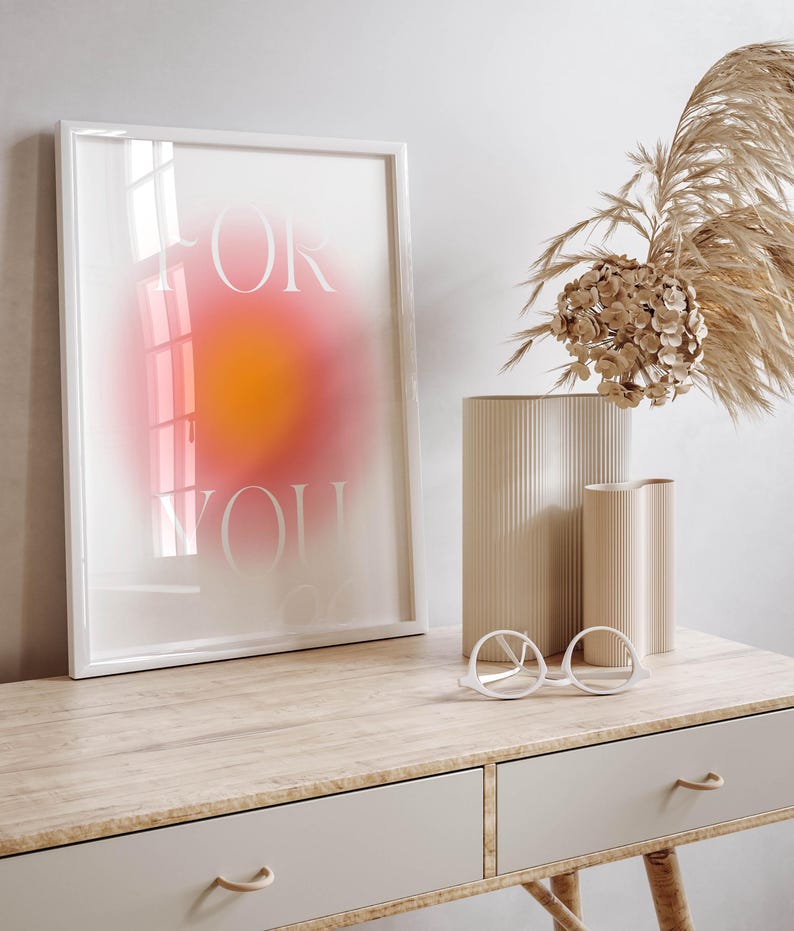 Peut inclure: Une impression encadr&eacute;e blanche avec les mots "FOR YOU" en police blanche sans empattement sur un fond d&eacute;grad&eacute; rose et orange. L'impression est pos&eacute;e sur un bureau en bois clair avec deux tiroirs et une paire de lunettes rondes blanches.