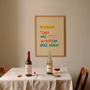 May include: A white tablecloth with two bottles of wine, one red and one white, and a glass of red wine. There is a plate with bread and a cork on the table. A framed picture with a five-star rating and the text "Chef was cute, would eat here again." is hanging on the wall behind the table.
