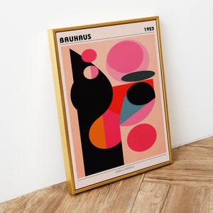 May include: A framed print featuring a geometric abstract design in pink, red, orange, black, and blue. The print includes the text "BAUHAUS" and "1923" in black lettering. The print is framed in a gold frame.