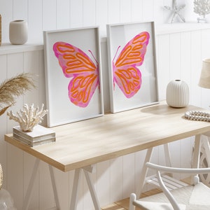 May include: Two white framed prints of pink and orange butterfly illustrations on a white wall above a light wood desk with a white chair, a vase, and a string of white beads.