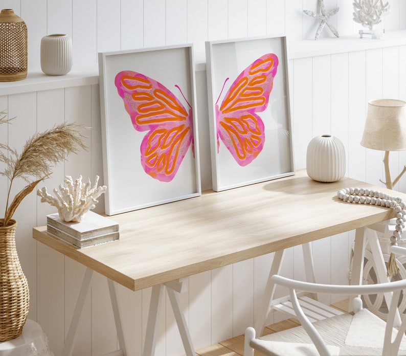 May include: Two white framed prints of pink and orange butterfly illustrations on a white wall above a light wood desk with a white chair, a vase, and a string of white beads.