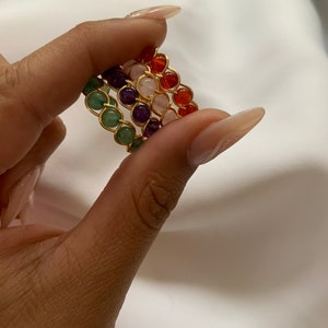 May include: Four stacked rings made of gold wire with different colored gemstones. The rings are green, purple, pink, and orange.