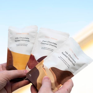 May include: Three individual pouches of chocolate-covered snacks. The pouches are white with brown and orange abstract designs. The pouches are labelled with the text "little.cocoa" and the flavour of the snack. The flavours are "Milk Chocolate Spiced Almonds", "Dark Chocolate Coconut Cherries", and "Milk Chocolate Roasted Hazelnuts".