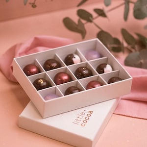 May include: A white cardboard box containing twelve assorted spherical chocolates with glossy finishes in shades of dark brown, metallic red, and pale purple. The box lid is placed underneath, featuring the text little. cocoa printed in brown.