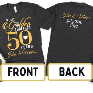 May include: A dark gray t-shirt with gold lettering that says "We are Golden Together 50 Years John & Maria". The back of the shirt has gold lettering that says "John & Maria July 21st, 1973".