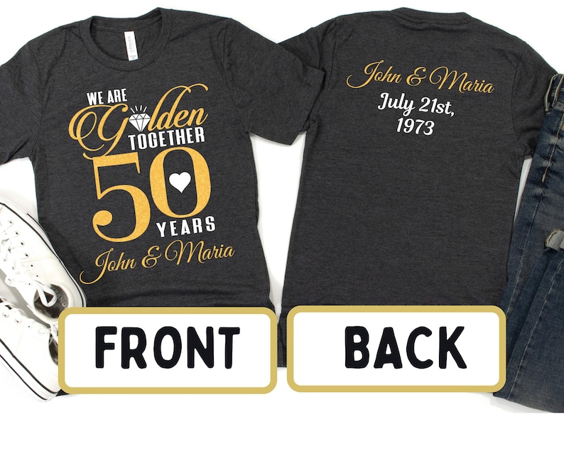 May include: A dark gray t-shirt with gold lettering that says "We are Golden Together 50 Years John & Maria". The back of the shirt has gold lettering that says "John & Maria July 21st, 1973".