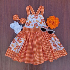 May include: Orange and white gingham dress with a white bodice featuring a pattern of cartoon girls. The dress has two pockets and a criss-cross strap design in the back.
