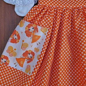 May include: Orange and white gingham fabric dress with a white pocket featuring a repeating pattern of butterflies and girls with red hair.
