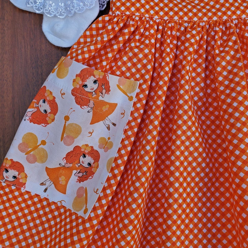 May include: Orange and white gingham fabric dress with a white pocket featuring a repeating pattern of butterflies and girls with red hair.